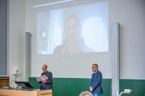 Zwei Professoren stehen vorn im Hörsaal. An der Wand ist eine Videoschalte projeziert.