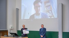 Der Dekan zeigt die Urkunde, während im Hintergrund der Preisträger in einer Videoschalte gezeigt wird.