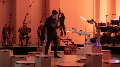 ein Dirigent steht neben drei Menschengroßen Roboterarmen, jeder mit einemTaktstock. Hinter der Szenerie sitzen vier Blechbläser eines Sinfonieorchersters.