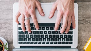 Das Foto zeigt von oben die Hände einer Person, welche eine Tastatur bedienen, daneben sind zur linken Seite eine Pflanze, sowie Kopfhörer und zur rechten Seite ein gelbes Buch.