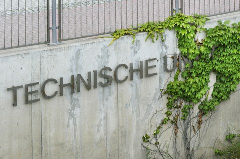 Das Foto zeigt eine Mauer an welcher der Schriftzug " Technische Universität" angebracht ist. Das Wort "Universität" ist von Efeu überwachsen.