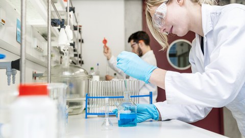 Photo shows two people working in the laboratory. They are wearing work clothes and protective goggles.