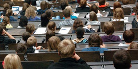 Das Foto zeigt einige Sitzreihen eines Hörsaals voller Studierender aus der Perspektive einer im Saal sitzenden Person. Viele der Studierenden schauen nach vorn oder machen sich Notizen.