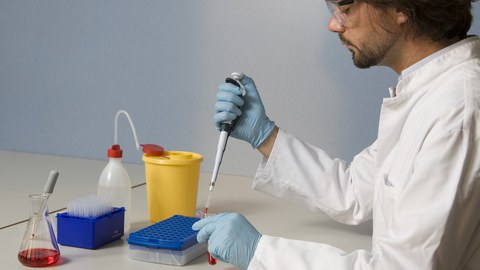 Das Foto zeigt eine Person, welche Arbeitskleidung und Schutzbrille tragend an einem Tisch sitzt, auf welchem verschiedene Laborutensilien stehen. Die Person füllt mit einer Spritze eine rote Flüssigkeit in ein Reagenzglas. 