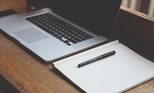Das Foto zeigt einen Laptop mit schwarzem Bildschirm, rechts daneben ein geöffnetes, leeres Notizbuch und einen daraufliegenden Stift  auf einer hölzernen Oberfläche.