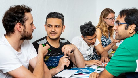 Foto von mehreren Personen verschiedener Nationen, die sich unterhalten und gemeinsam Aufgaben lösen.