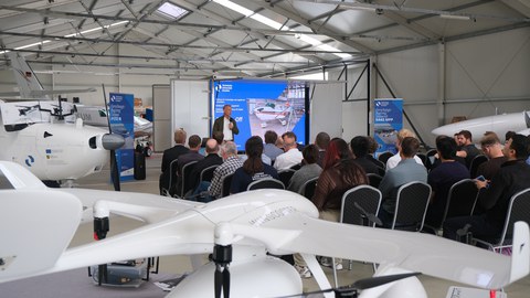 Teilnehmende einer Forschungsdemonstration in einem Flugzeughangar; im Vordergrund eine weiße VTOL-Drohne, im Hintergrund hält ein Referent einen Vortrag vor einer Leinwand.