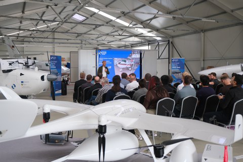 Participants attending a research demonstration inside an aircraft hangar, with a white VTOL drone in the foreground and a presenter speaking in front of a screen.