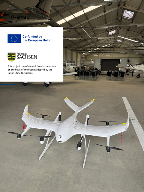 White VTOL research drone with multiple rotors and fixed wings inside a hangar, with several crewed aircraft visible in the background.