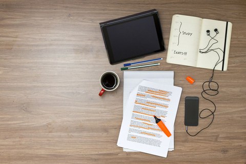 Das Foto zeigt mehrere Gegenstände auf einem Tisch. Man erkennt ein Tablet, mehrere Stifte, einige Zettel, ein Notizbuch, ein Handy und eine Tasse Kaffee.