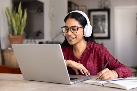 Smiling student woman wearing wireless headphones studying online sits at table at home using laptop and notebook