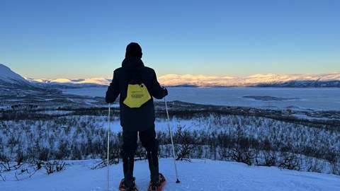 Im Vordergrund steht eine Person mit dem Rücken zu uns und schaut auf eine verschneite Landschaft herab. 