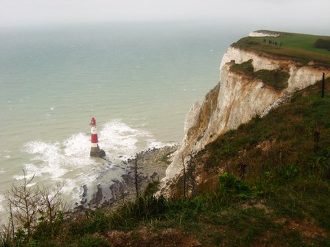 white chalk cliffs overlooking the sea. At the beach down below, a lighthouse with red and white stripes is standing between the waves