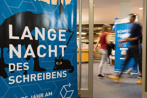 Im Foyer der SLUB-Zweigbibliothek Erziehungswissenschaft stehen zwei Roll-Up-Banner: Auf einem steht "Lange Nacht des Schreibens", auf dem anderen "Schreibzentrum". Zwei Personen laufen durch das Bild, sind aber durch ihre Laufbewegung nicht erkennbar.