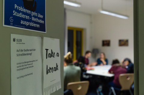 Ein Blick an der halb geöffneten Tür vorbei in den Raum der Schreibgruppe "Probieren geht über Studieren - Methoden ausprobieren": Die Teilnehmenden sitzen an Tischen und sprechen miteinander.