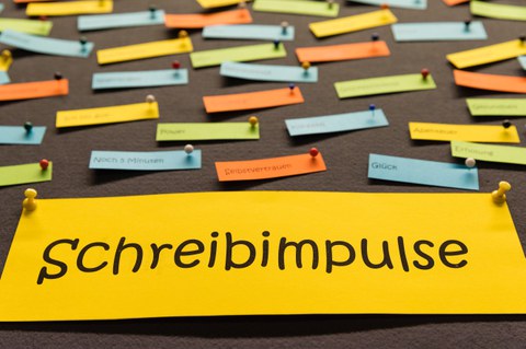 An eine Pinnwand sind viele verschiedenfarbige Zettel geheftet. Auf einem großen gelben Zettel steht "Schreibimpulse".