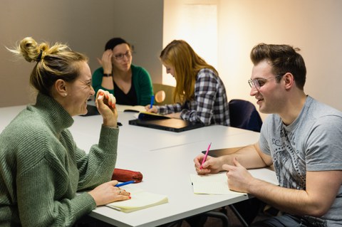 Vier Studierende sitzen an einem Tisch. Sie haben jeweils einen Block und einen Stift vor sich und sprechen in Zweiergruppen miteinander.