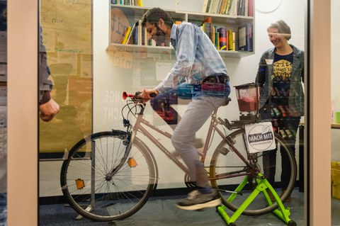 Hinter einer Glasscheibe steht ein aufgeFahrrad, auf dessen Gepäckträger ein Mixer montiert ist. Ein Mann sitzt auf dem Rad
