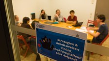 Durch die Glasschreibe eines Gruppenarbeitsraumes sieht man eine Gruppe Studierender. An der Tür hängt ein Schild mit dem Titel der Schreibgruppe: "Strategien & Arbeitsweisen finden: strukturschaffend oder -folgend?