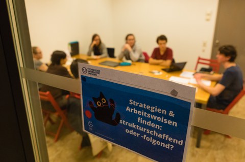 Durch die Glasschreibe eines Gruppenarbeitsraumes sieht man eine Gruppe Studierender. An der Tür hängt ein Schild mit dem Titel der Schreibgruppe: "Strategien & Arbeitsweisen finden: strukturschaffend oder -folgend?