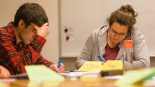 Zwei Studierende sitzen mit gesenkten Köpfen und konzentrierten Blicken über ihren Arbeitspapieren.