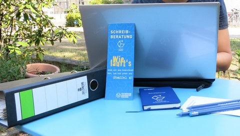 Auf einem Tisch steht ein aufgeklappter Laptop, davor lehnt ein Lesezeichen mit "Schreibberatung! Läuft bei dir?", rechts daneben liegen Bleistifte, links daneben ein Ordner.