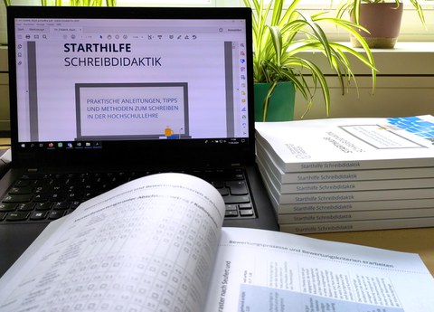 Ein Laptop mit dem geöffneten PDF der Starthilfe Schreibdidaktik, davor das geöffnete Printexemplar und daneben ein Stabel geschlossener Printexemplare.