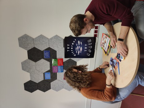 Zwei Personen sitzen an einem runden Tisch, blicken auf Zeitungen und Rätsel auf dem Tisch. Im Hintergrund: Pinnwand mit Postkarten und ein Poster mit dem Text: "Try different. Try again."