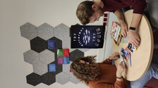 Zwei Personen sitzen an einem runden Tisch, blicken auf Zeitungen und Rätsel auf dem Tisch. Im Hintergrund: Pinnwand mit Postkarten und ein Poster mit dem Text: "Try different. Try again."
