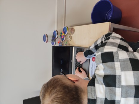 Ein Tisch mit einem Laptop mit schwarzem Bildschirm, davor ein Mann in kariertem Hemd, der auf der Tastatur tippt. Hinter dem Laptop: zu einer Pyramide gestapelte Schraubgläser mit unterschiedlichem Inhalt: Büroklammern, Papierrollen, Pins, Tintenpatronen, Zahnzwischenraumreiniger.