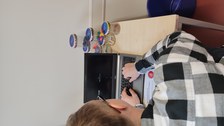 Ein Tisch mit einem Laptop mit schwarzem Bildschirm, davor ein Mann in kariertem Hemd, der auf der Tastatur tippt. Hinter dem Laptop: zu einer Pyramide gestapelte Schraubgläser mit unterschiedlichem Inhalt: Büroklammern, Papierrollen, Pins, Tintenpatronen, Zahnzwischenraumreiniger.