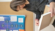 Eine Frau sitzt auf einem Stuhl mit einer geöffneten Holzschatulle in der Hand. In der Schatulle: puzzleartige Holzteile. Dahinter: Pinnwand mit einem Poster eines Ablaufschemas, Inhalt nicht erkennbar.