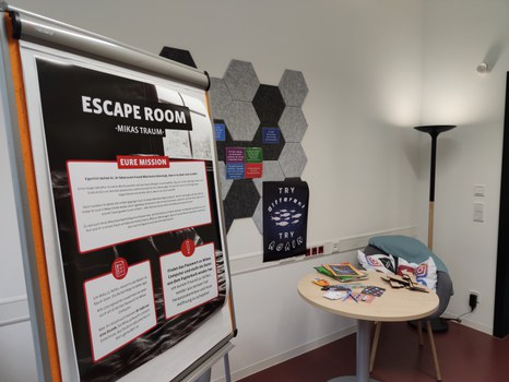 Teil eines Raumes, im Vordergrund: Plakat "Escape Room - Mikas Traum" mit Informationskästen "Deine Mission" und Hinweisen, die nicht lesbar sind. Dahinter Postkarten auf einer Pinnwand, ein Tisch mit Zeitungen, Stiften und Rätseln, ein Sessel mit Kissen.