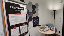 Teil eines Raumes, im Vordergrund: Plakat "Escape Room - Mikas Traum" mit Informationskästen "Deine Mission" und Hinweisen, die nicht lesbar sind. Dahinter Postkarten auf einer Pinnwand, ein Tisch mit Zeitungen, Stiften und Rätseln, ein Sessel mit Kissen.