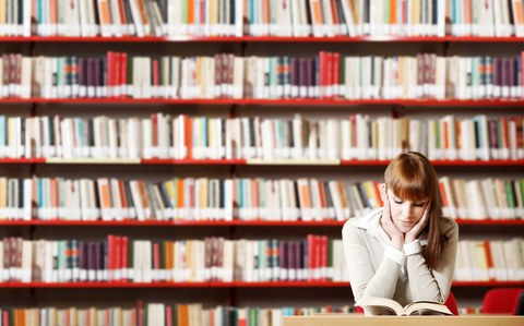 Photo of a young student sitting at a table in front of a wall of books and reading a book.