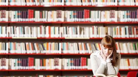 Photo of a young student sitting at a table in front of a wall of books and reading a book.