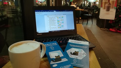 Ein Arbeitsplatz in einem bekannten Coffeeshop, zu sehen ist ein geöffneter Laptop, darauf Flyer des Schreibzentrums, links eine Tasse Kaffee, im Hintergrund Cafébesucher.