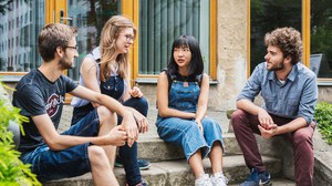 Auf einer Treppe sitzen vir Studierende. Außen sitzen zwei Männer, in der Mitte sitzen zwei Frauen. Die Studierenden unterhalten sich miteinander. 
