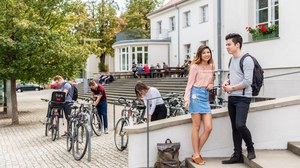Zwei Studierende stehen auf der Treppe vor der Mensa und unterhalten sich. Im Hintergrund stehen weitere Studierende. 