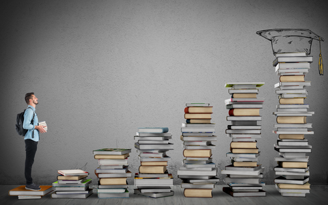 Man with books in hands and backpack on his back stands on the first step of a staircase, consisting of individual stacks of books, with an alumni hat on the top stack
