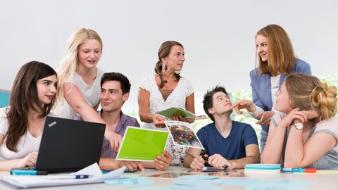 Auf diesem Foto sind mehrere Personen während eines Workshops zu sehen. Fünf Studierende sitzen an einem großen Tisch, auf welchem unterschiedliche Büroutensilien liegen. Zwei Frauen stehen am Tisch und wenden sich den Studierenden zu. 