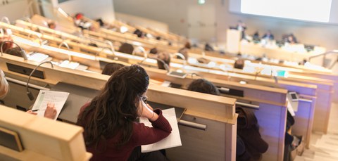 Das Foto zeigt eine Studentin im Hörsaal. Sie stützt ihren Kopf auf Ihre Unterlagen, die vor ihr auf dem Tisch liegen.