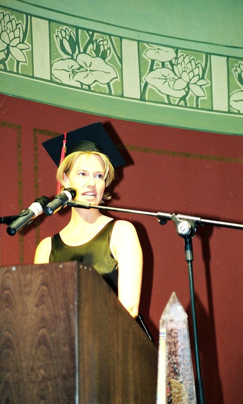 The photo depicts Annikka Zurwehme with a graduation cap standing at a podium with a microphone.