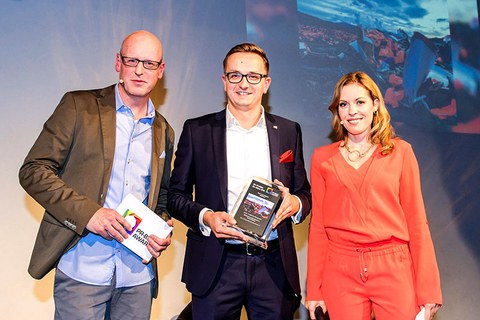The photo shows three people at an award ceremony. Kai Kranich stands in the center showing the prize to the camera.