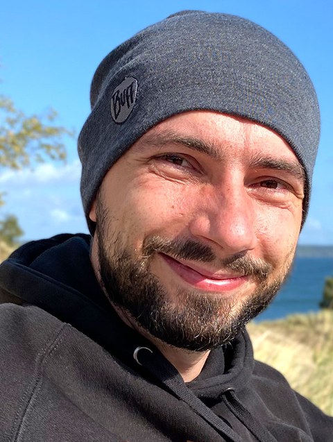 Photo portrait of Carsten Bittrich outside. He is wearing a hat and there is a body of water in the background.