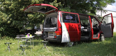 Das Foto zeigt einen Campingvan im Grünen. Davor steht ein Tisch mit Stühlen.