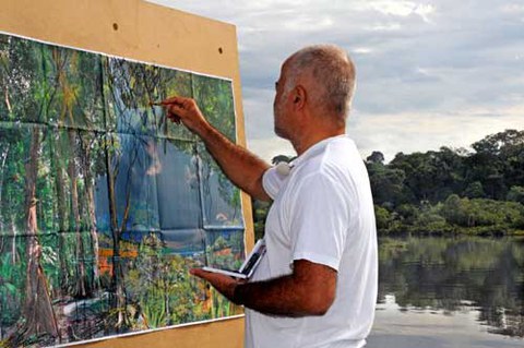 The photo shows Yadegar Asisi standing before and painting on a canvas. In the background are trees and a body of water.