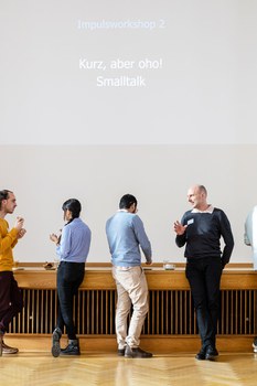 Teilnehmende stehen in Zweiergruppen vor der Bühne mit Präsentation