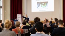 Teilnehmende sitzen vor der Bühne und einige heben die Hand. Moderatorin steht vor der Bühneund hebt die Hand