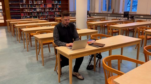 Ein Foto von einem Lesesaal in einer Bibliothek. Im Hintergrund ist ein Regal mit Büchern zu sehen. Davor sind Tische angeordnet. Mittig sitzt ein Mann mit kurzem dunklem Haar an einem der Tische und schaut in seinen Laptop.